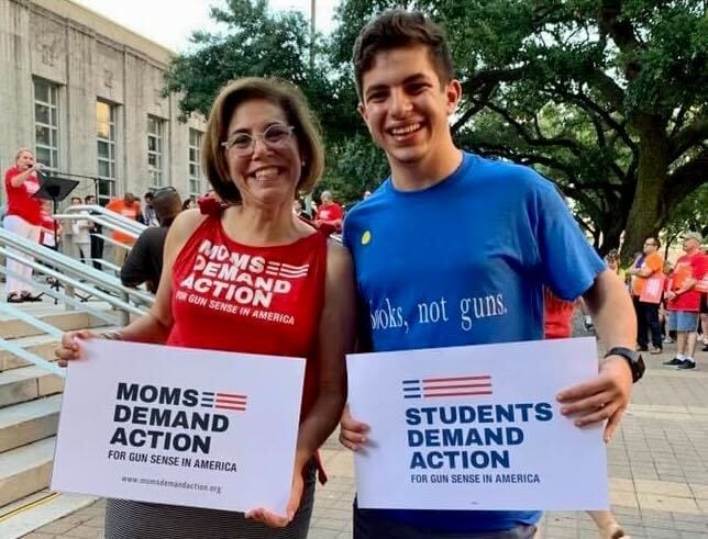 Norri smiles while holding a Moms Demand Action sign next to a person holding a Students Demand Action sign 