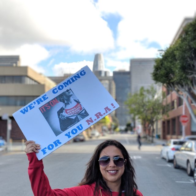 CA volunteer Alex Navarrro smiles while wearing a red Moms Demand Action shirt and holding up a sign that says 'we're coming for you NRA!'
