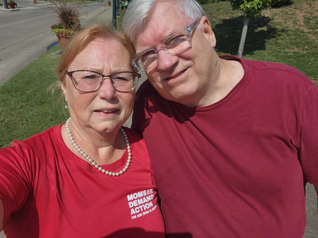 A white husband and wife take a selfie while canvassing. The wife wears a red Moms Demand Action t-shirt, wire-rimmed oval glasses, and a pearl necklace. Her husband wears a plain burgundy t-shirt and rectangular glasses with a clear frame.