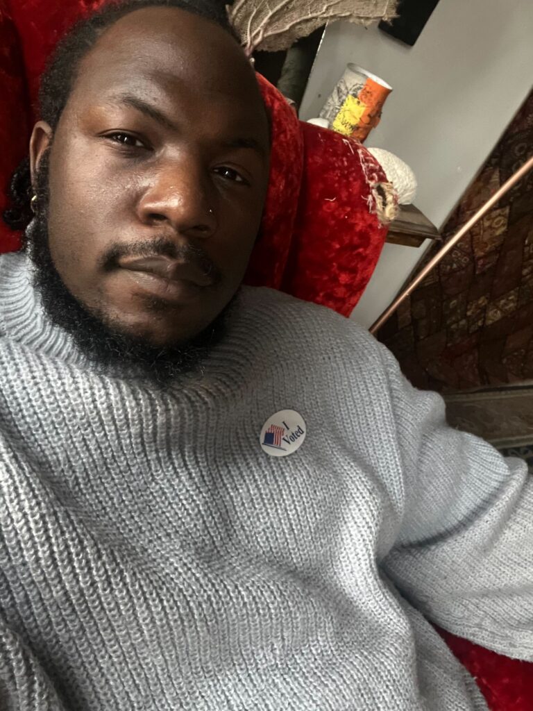 A Black gun violence survivor takes a selfie. He is wearing a grey ombre turtleneck sweater and an "I Voted" sticker.