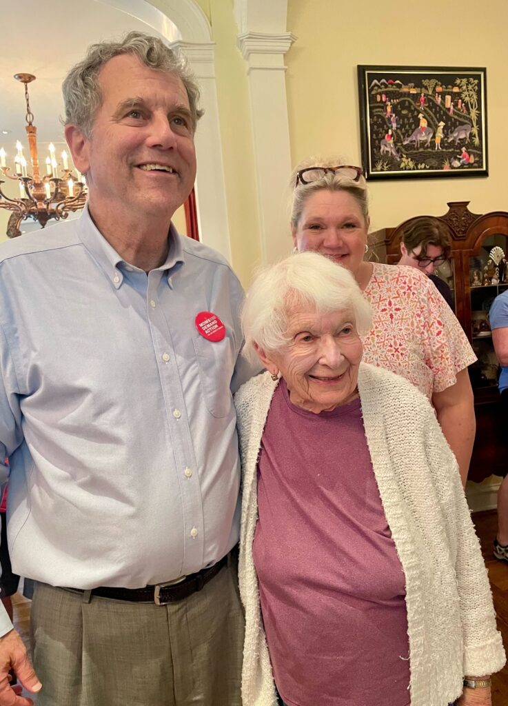 A 97-year-old white woman with white hair poses for a photo with Sherrod Brown. The woman wears a burgundy t-shirt and a white knit cardigan. Sherrod is a white man and wears a blue button-up shirt, grey dress pants, and a red Moms Demand Action button on his shirt pocket.