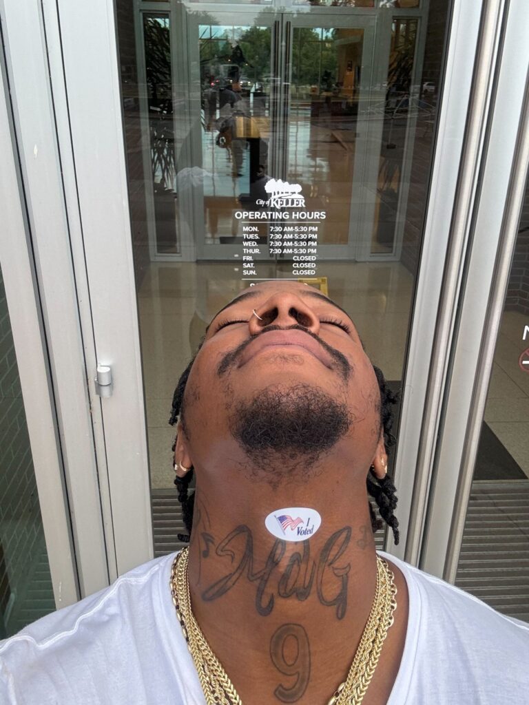 A Black gun violence survivor takes a selfie at his polling place. His head is tipped back and he has placed his "I Voted" sticker on his throat above his Adam's apple; directly below the sticker, he has a tattoo that appears to read "MdG" with the number "9" underneath it.