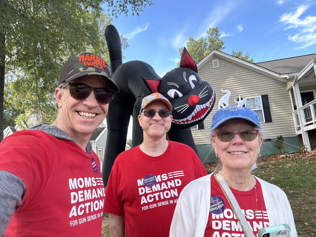 Three white volunteers wear red Moms Demand Action t-shirts. They are standing in front of a house with a giant black inflatable cat Halloween decoration in the yard.