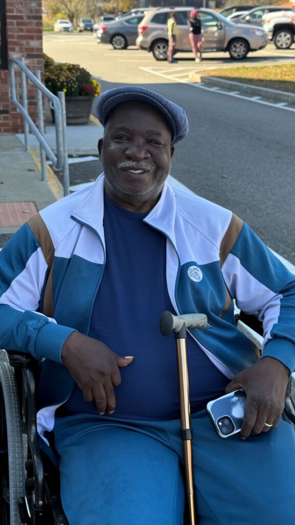 A Black gun violence survivor has his photo taken after voting at his polling place. He is sitting in a wheelchair and has a cane propped against his legs. He holds his phone in his left hand, has a blue/grey newsboy-style cap, and wears a white-and-blue jacket.