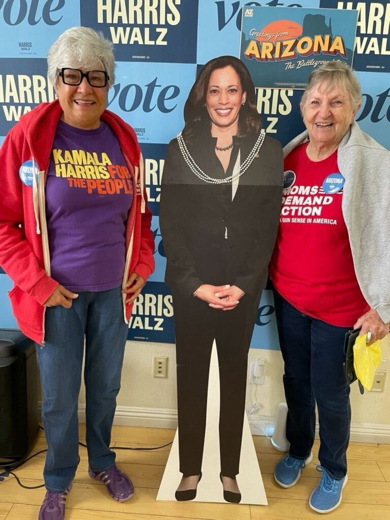 Two Moms Demand Action volunteers stand with a cardboard cutout of Kamala Harris, who is wearing a black suit, in between them. The cutout has a necklace with three strings of pearls draped around Kamala's neck. One volunteer wears a red Moms Demand Action sweatshirt and a purple t-shirt that says "Kamala Harris: For the People." The other volunteer wears a red Moms Demand Action t-shirt.