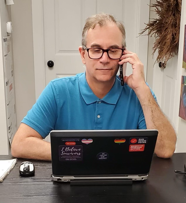 A white man phonebanks; he is holding his phone up to his left ear while staring at his laptop. The laptop is covered with stickers that read "Gun Sense Voter", "Moms Demand Action", "We Can End Gun Violence", and "I believe survivors". He is wearing a blue polo shirt and dark rectangular glasses.