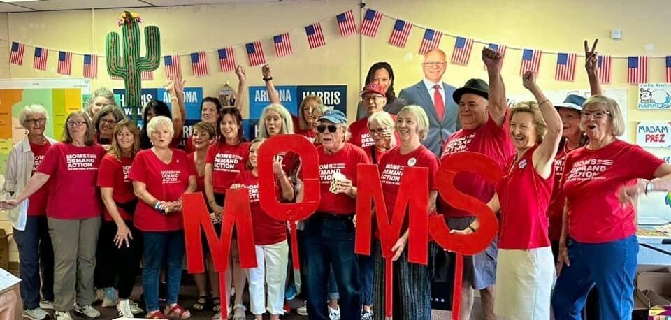 Moms Demand Action volunteers in Tucson, Arizona, wear red Moms Demand Action t-shirts. In the back of the group, you can see cardboard cutouts of Kamala Harris and Tim Walz. A fake cactus is propped against the back wall; a string banner with small American flags is hanging on the back wall.