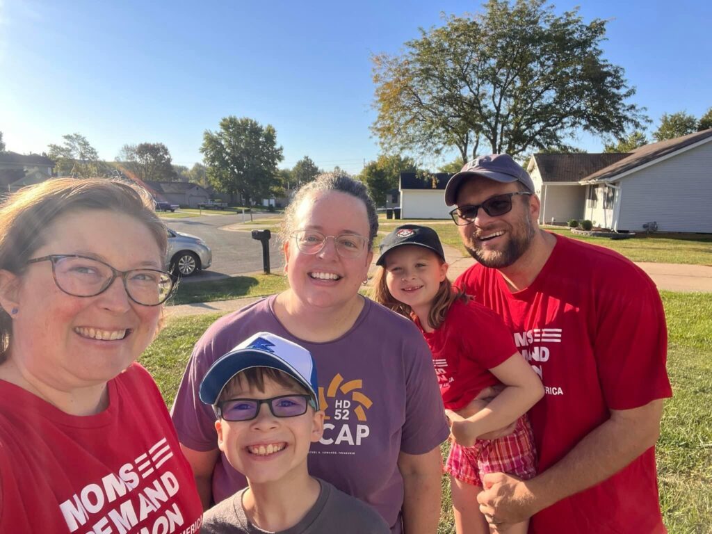 Three adults and two children take a selfie. Two of the adults and one of the children are wearing red Moms Demand Action t-shirts. A suburban streets is visible in the background.