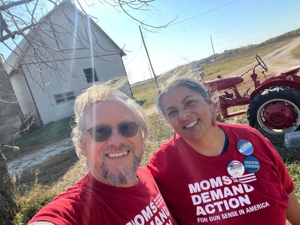 A husband and wife take a photo while canvassing. A barn and a tractor are visible behind them. They both wear red Moms Demand Action t-shirts.