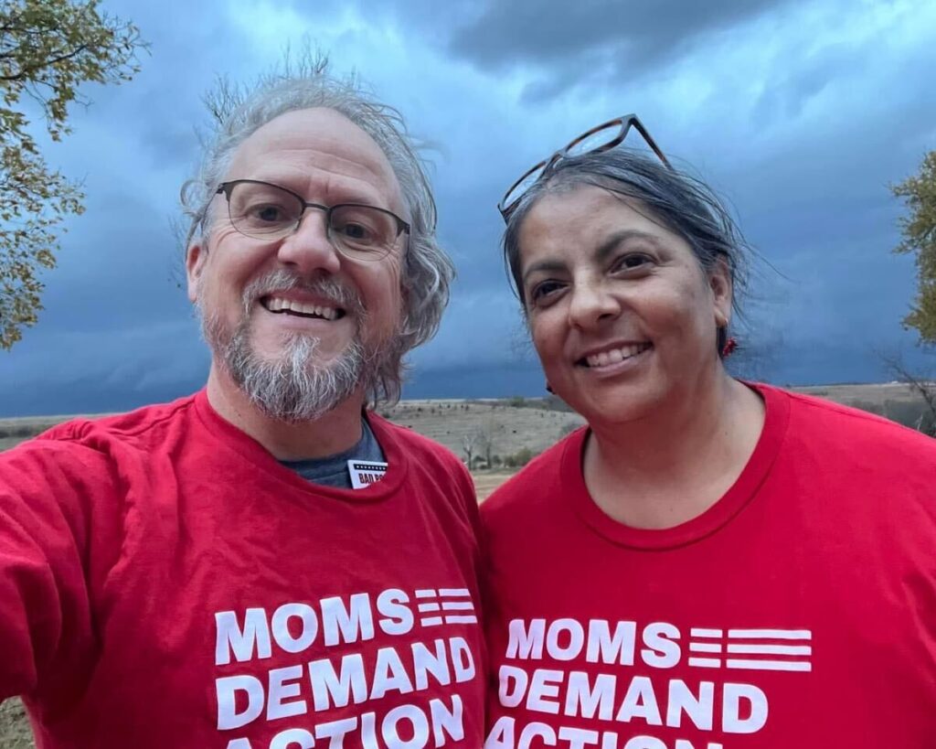 A husband and wife take a selfie. They are wearing red Moms Demand Action t-shirts. The sky behind them is dark and seems to indicate that a storm is brewing; farmland is in the background.