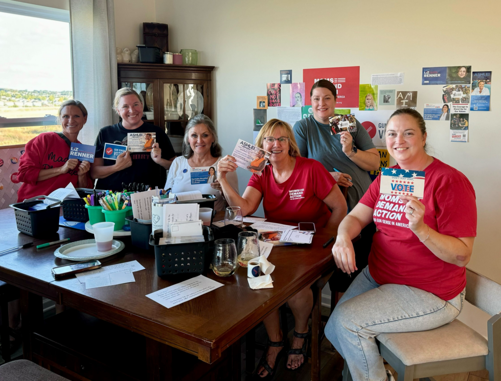 Six Moms Demand Action volunteers sit around a wood table and smile for the camera. They are all holding up postcards that they have written to support Gun Sense Candidates.