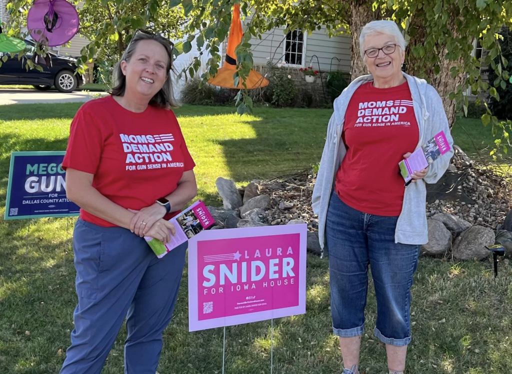 Two Moms Demand Action volunteers pose for a photo in front of a candidate yard sign. They are both white and are wearing jeans and red Moms Demand Action t-shirts.