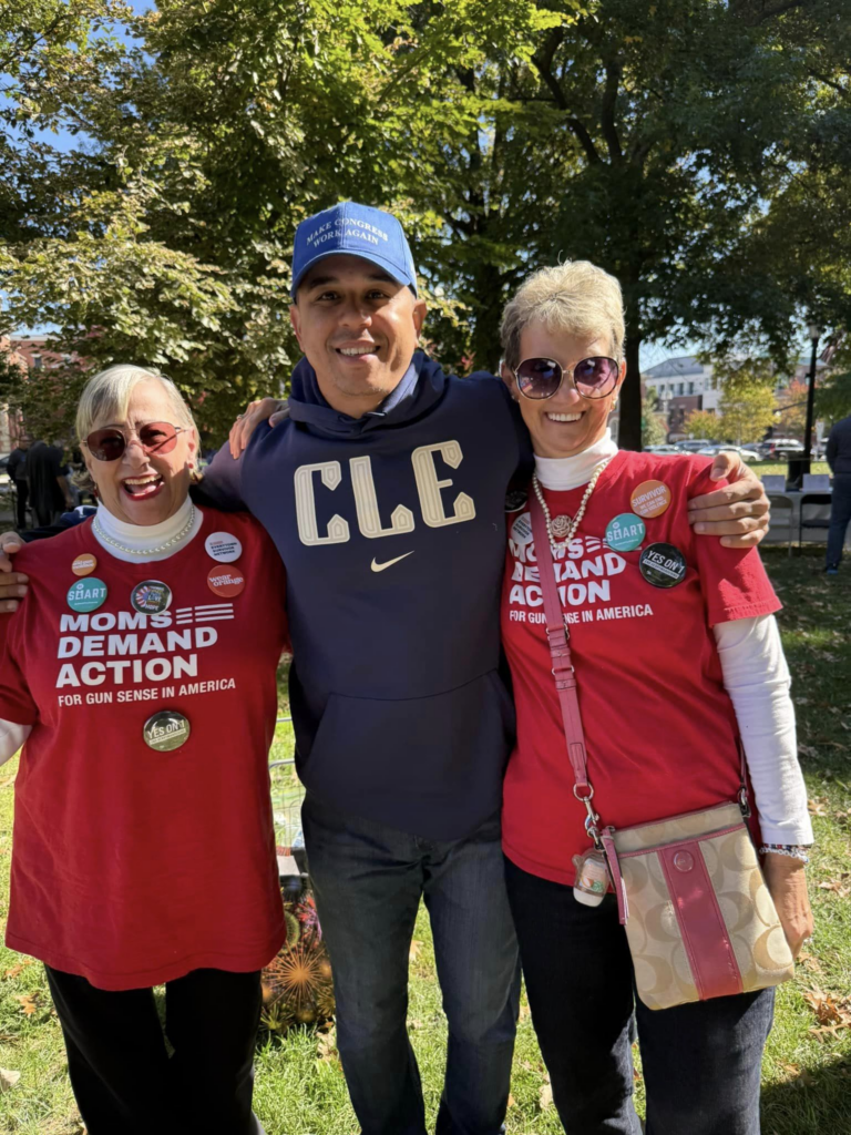 Two Moms Demand Action volunteers pose with a Cleveland Browns baseball player standing in between them. He is wearing a navy blue sweatshirt with yellow "CLE" letters and a Nike swoosh on the chest. He is wearing a blue baseball cap that reads "Make Congress Boring Again." Both volunteers are wearing dark sunglasses and red Moms Demand Action t-shirts over top of white turtleneck sweaters with various buttons.