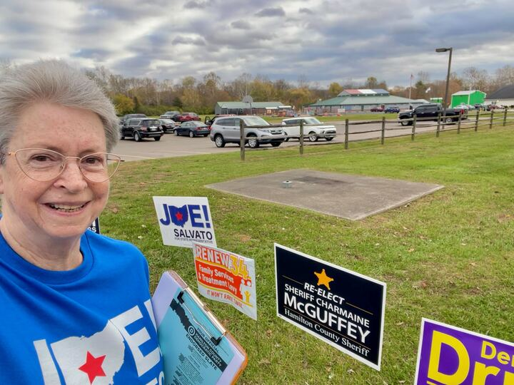 A white woman with grey short hair, gold wire-rimmed classes, and a blue t-shirt takes a selfie in front of several political signs at a poling place. She has voting slates attached to a clipboard tucked under her left arm.