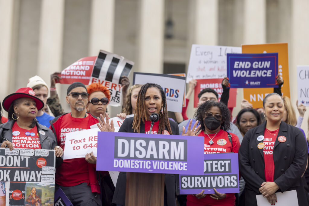 Angela Ferrell-Zabala, the executive director of Moms Demand Action, speaks at a "disarm domestic violence" rally in front of the Supreme Court in November 2023 prior to oral argument in the United States v. Rahimi Supreme Court case. She is standing at a podium with a crowd of supporters behind her wearing Moms Demand Action t-shirts and holding signs advocating for gun violence prevention.