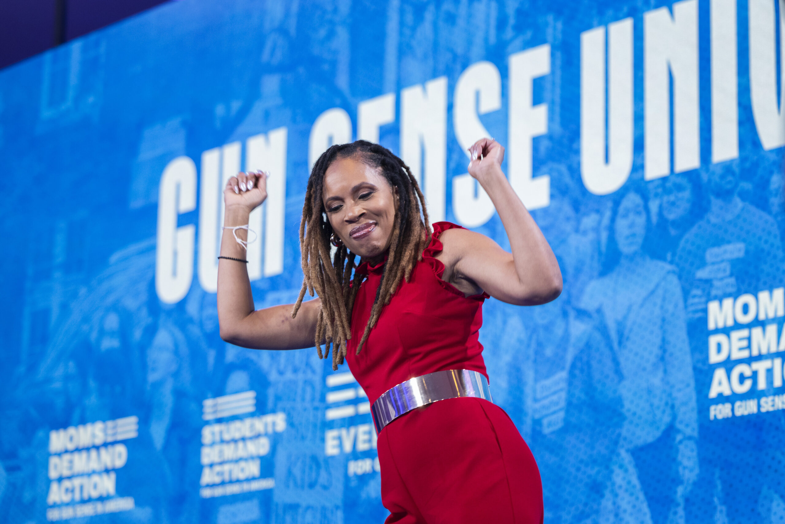 Angela Ferrell-Zabala, executive director of Moms Demand Action, is wearing a red jumpsuit with a metallic silver belt. She is dancing on stage with her arms raised at Gun Sense University in D.C. in 2024.