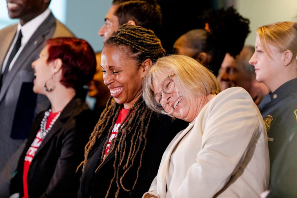 Angela Ferrell-Zabala, executive director of Moms Demand Action, sits at a bill signing in Michigan. She is laughing with another Moms Demand Action volunteer, who leans into her and rests her head on Angela's shoulder.