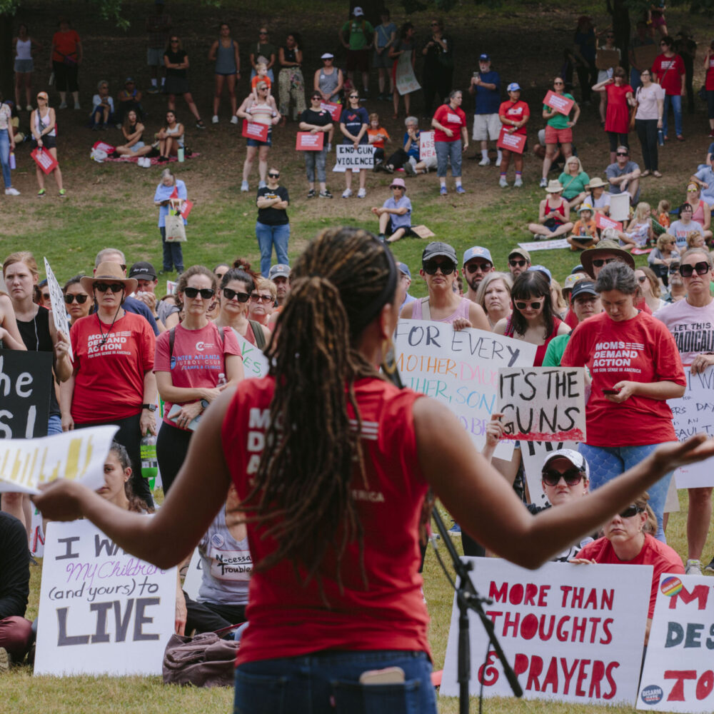Angela Ferrell-Zabala, executive director of Moms Demand Action, is pictured from behind while speaking to a crowd in Atlanta, Georgia. Members of the crowd hold signs saying things like