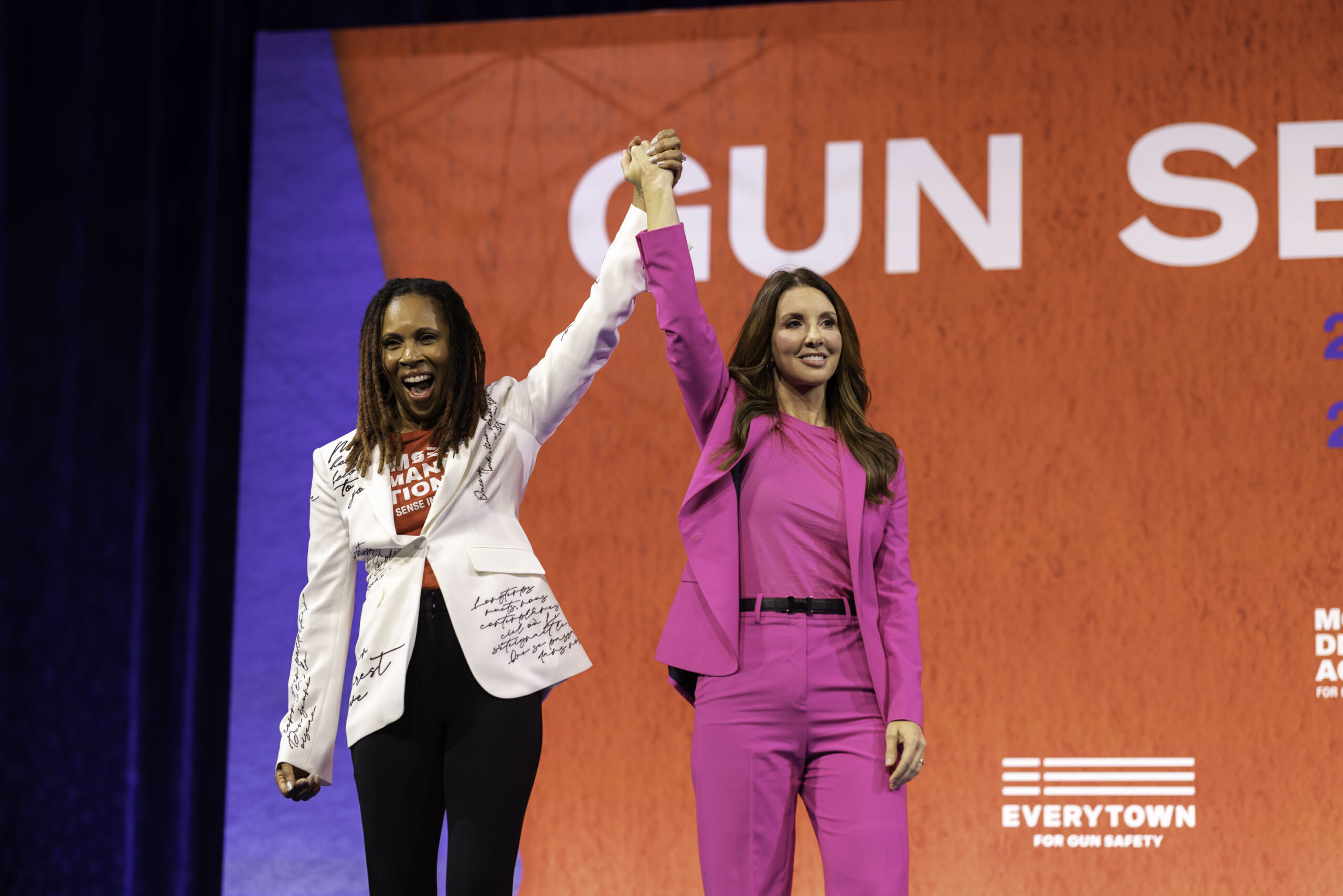 Angela Ferrell-Zabala, executive director of Moms Demand Action, links hands, raises arms, and smiles on stage at Gun Sense University in 2023 in Chicago with Shannon Watts, the founder of Moms Demand Action. Angela wears black pants, a white blazer, and a red Moms Demand Action t-shirt; Shannon wears a fuchsia pantsuit with a black belt.