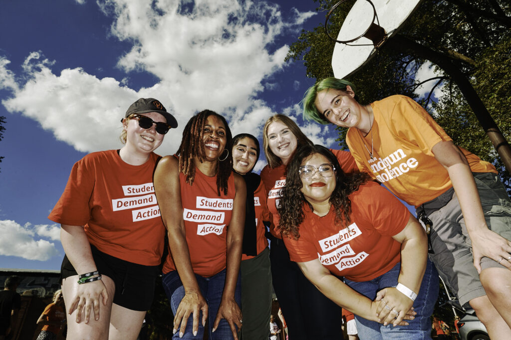 Angela Ferrell-Zabala, the executive director of Moms Demand Action, poses for a photo with five Students Demand Action volunteers. They are all smiling and slightly squatting down as they look at the camera, which is propped at a lower angle. They are outside at a basketball court that they are working to restore.