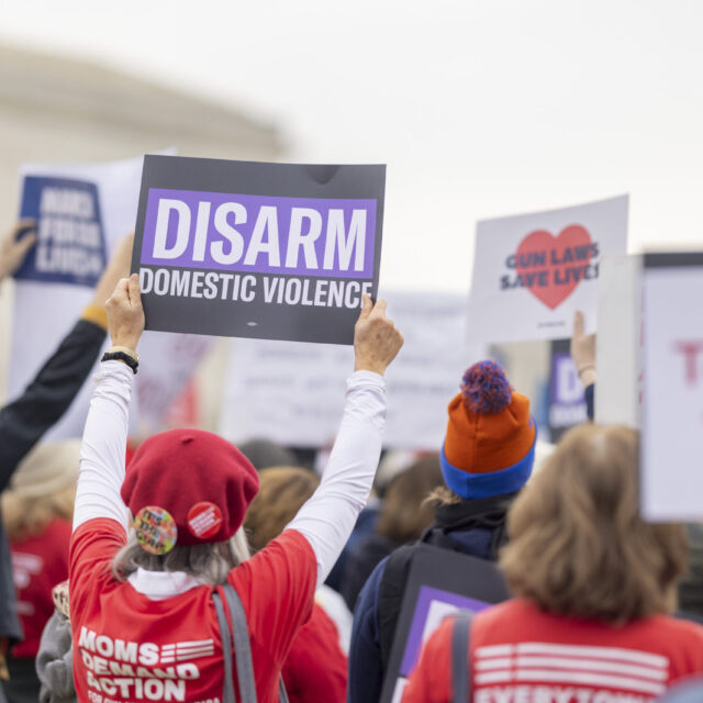 A view from the back of a crowd of Moms Demand Action volunteers protesting in front of the Supreme Court. One volunteer holds a sign that reads