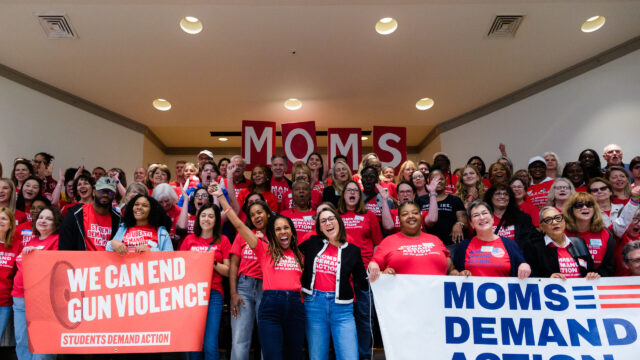A large group of people, all wearing red Moms Demand Action or Students Demand Action t-shirts, are smiling, clapping, and cheering in a group photo. In the front left, a group of people hold a banner that says 