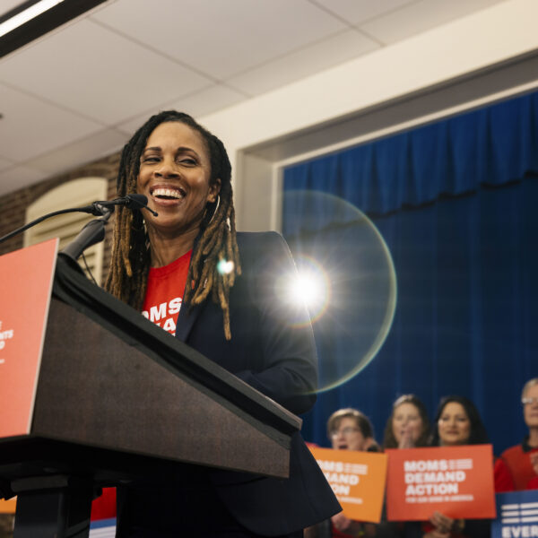 Angela Ferrell-Zabala, the executive director of Moms Demand Action and Students Demand Action, smiles while speaking at a podium. She wear metal hoop earrings, a black blazer, and a red Moms Demand Action t-shirt. Behind her, a cluster of 7 volunteers hold Moms Demand Action and Everytown placards and clap as she talks.