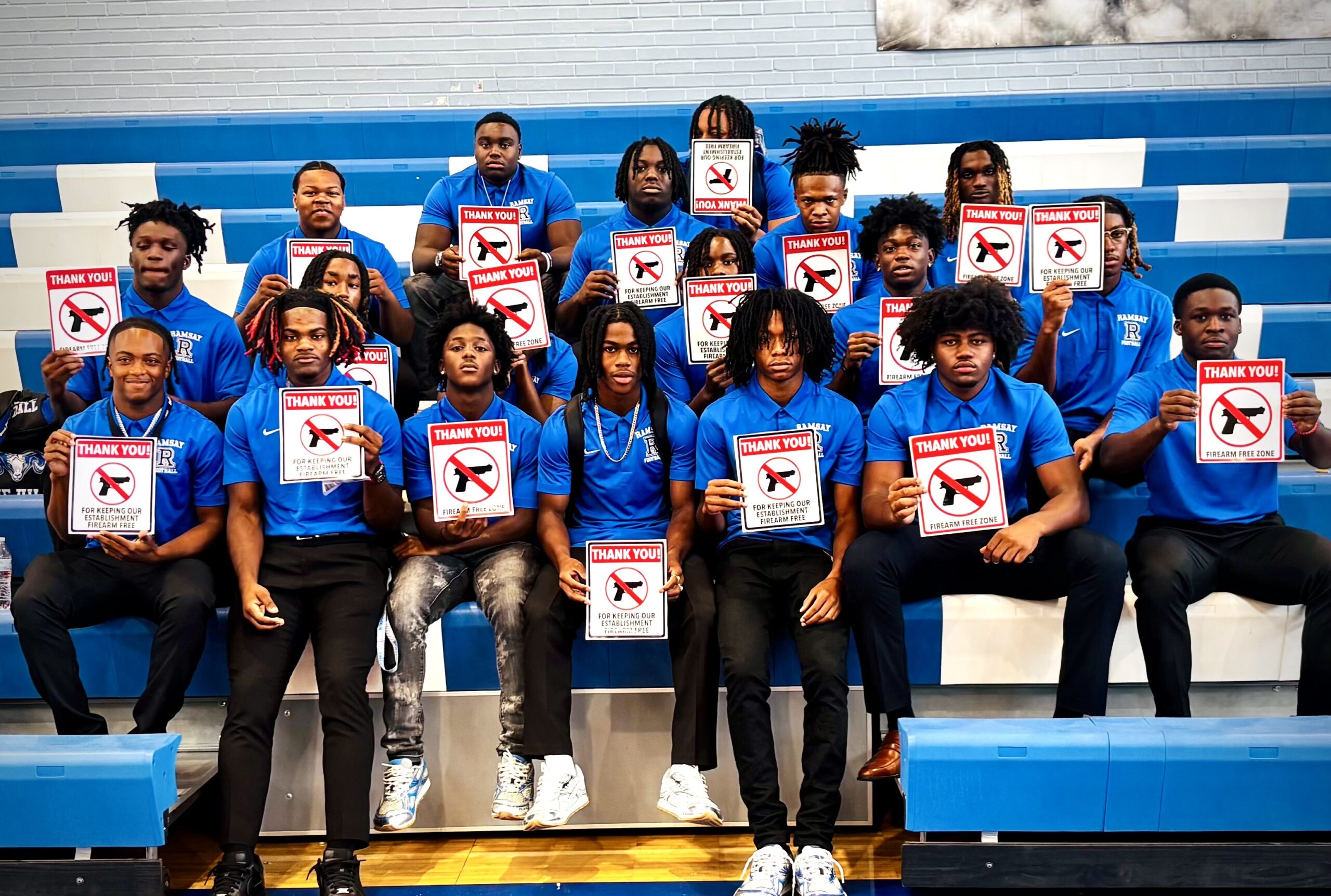 Members of the Ramsay High School football team sit in the bleachers at a pep rally and wear blue polo shirts, black pants, and hold signs that read "Thank you for keeping our establishment firearm free."