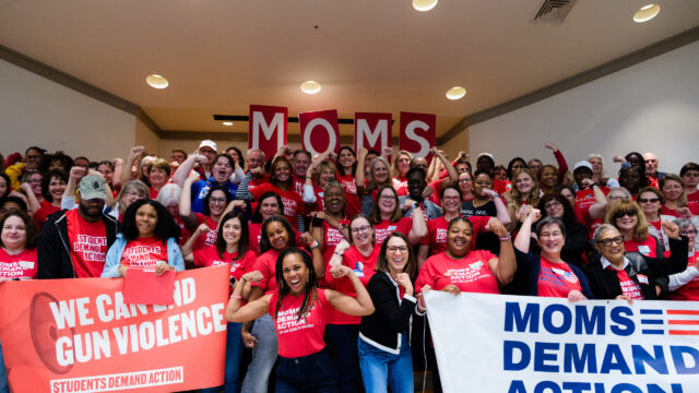 A group of over 100 Moms Demand Action and Students Demand Action volunteers, all wearing red shirts, pose for a photo. Most are holding one arm up and flexing their bicep. Angela Ferrell-Zabala, executive director of Moms Demand Action and Students Demand Action, has her sleeves rolled up and is flexing in between two groups of volunteers who are holding 