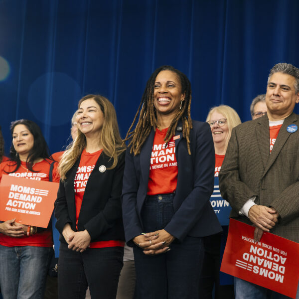 Moms Demand Action volunteers wear red t-shirts and blazers, holding Moms Demand Action signs, as they smile on stage at a rally. Angela Ferrell-Zabala, executive director of Moms Demand Action, smiles at the center of the photo.