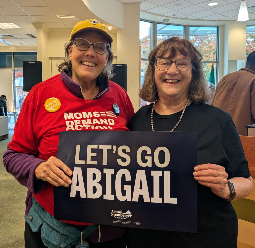 Two Moms Demand Action volunteers smile for a photo. They are holding up a sign that reads, "Let's Go Abigail" in capital letters.