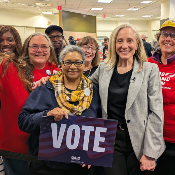 6 Moms Demand Action volunteers stand with Abigail Spanberger, who wears a black shirt, dark pants, and a light grey blazer. One volunteer holds a sign that says 