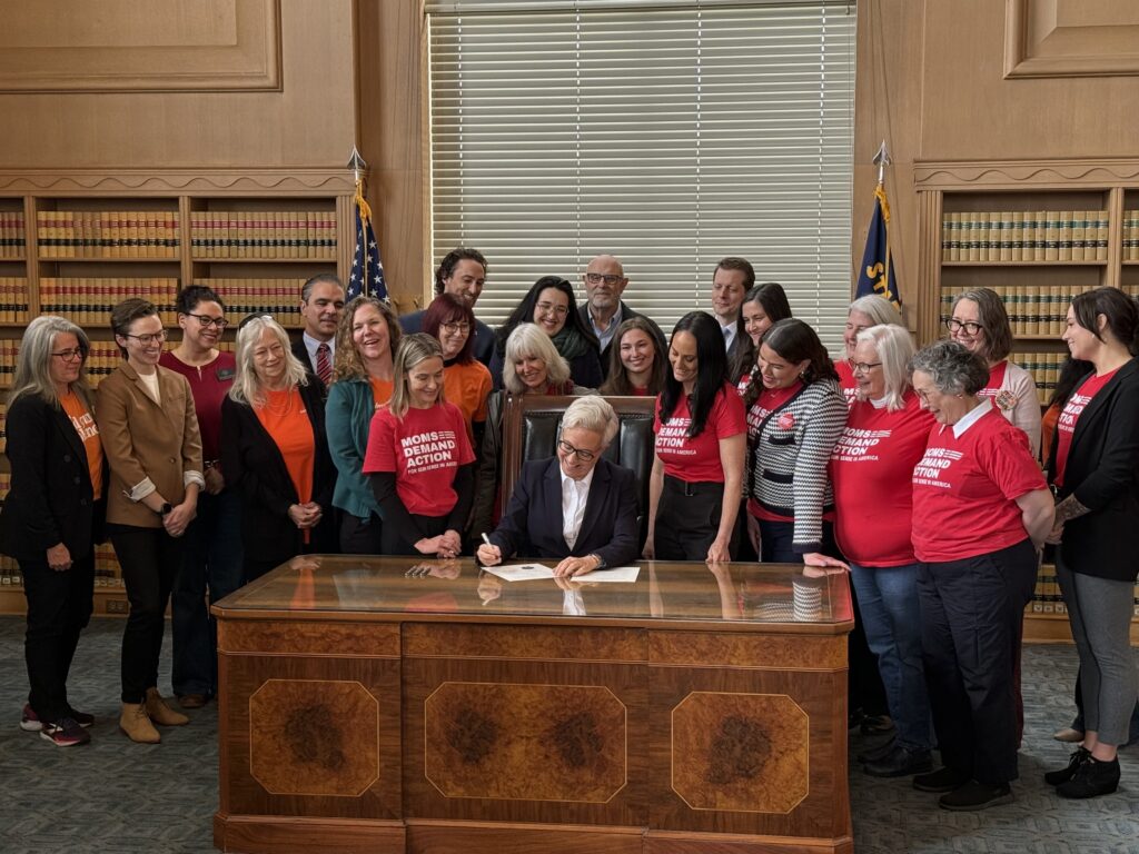 Governor Tina Kotek of Oregon sits at a wooden desk and looks down at a bill; she holds a pen in her right hand. She is surrounded by Moms Demand Action volunteers in red t-shirts and professional clothing, who are watching her sign the bill.